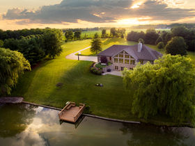 Aerial view of a luxury home surrounded by a manicured lawn, private dock on a tranquil pond, and a sunset sky, highlighting the property's stunning features and idyllic setting.