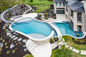 Aerial view of a luxury backyard with an infinity-edge pool, integrated hot tub, stone patio, landscaped rock garden, and a sleek water slide, adjacent to a modern home with large windows. Keywords: luxury backyard, infinity pool, high-end real estate, outdoor living space, aerial photography.