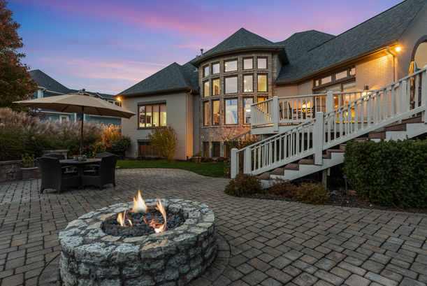 A backyard with a stone fire pit, paver patio, outdoor dining table with umbrella, a staircase to a deck, and a sunset-lit home with large windows.