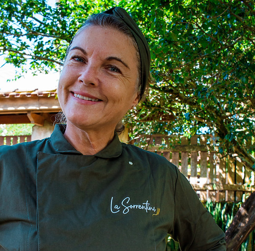 Chef da La Sorrentina sorrindo, vestindo uniforme verde com avental branco, em um ambiente ao ar livre cercado por árvores