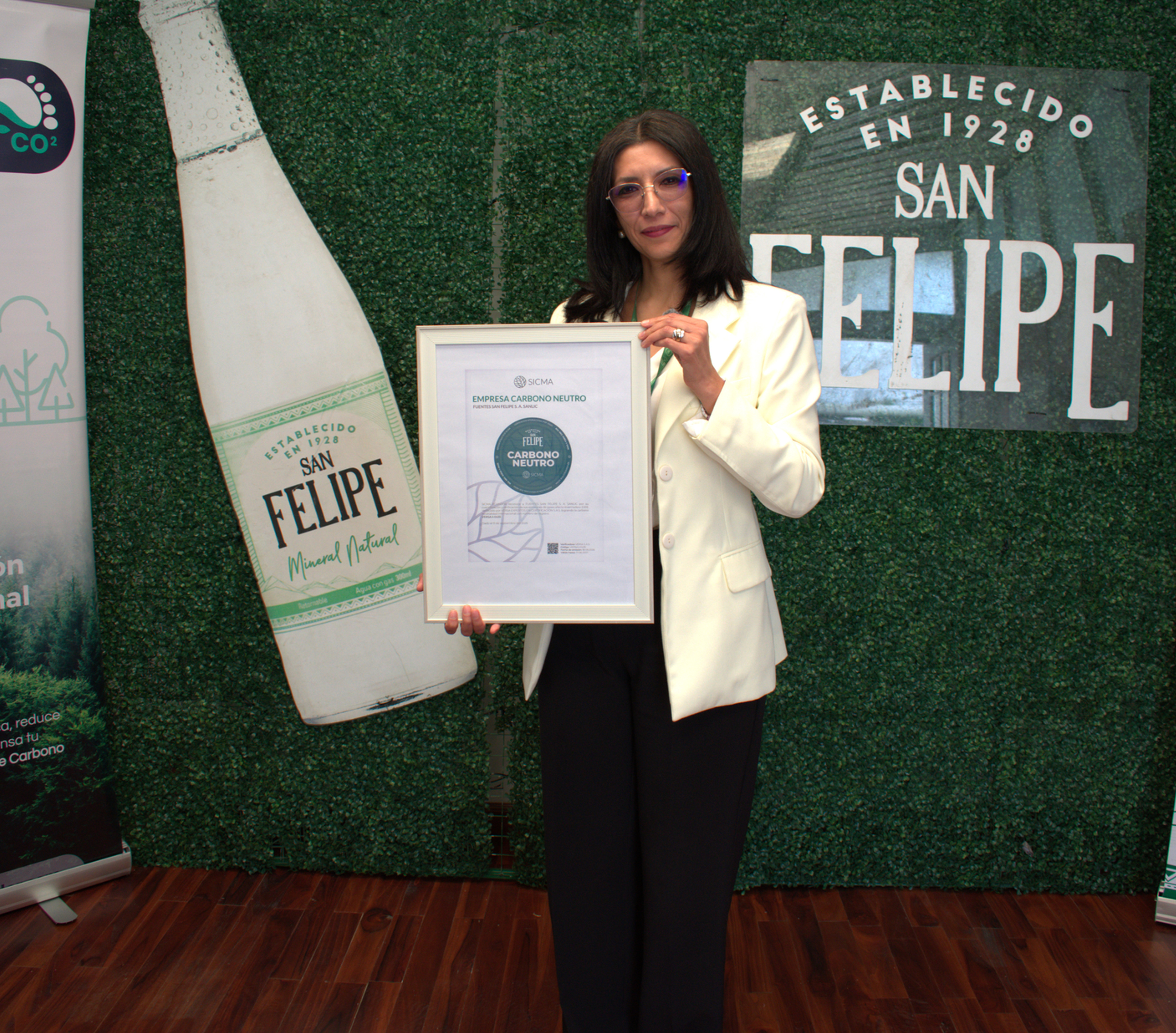 Mujer con blazer blanco sostiene un certificado en un evento de San Felipe con banners de carbono neutro.