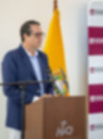 Hombre con gafas en podio, hablando ante micrófono. Fondo con bandera amarilla y logotipos de Hospital Vozandes Quito. Expresión seria.