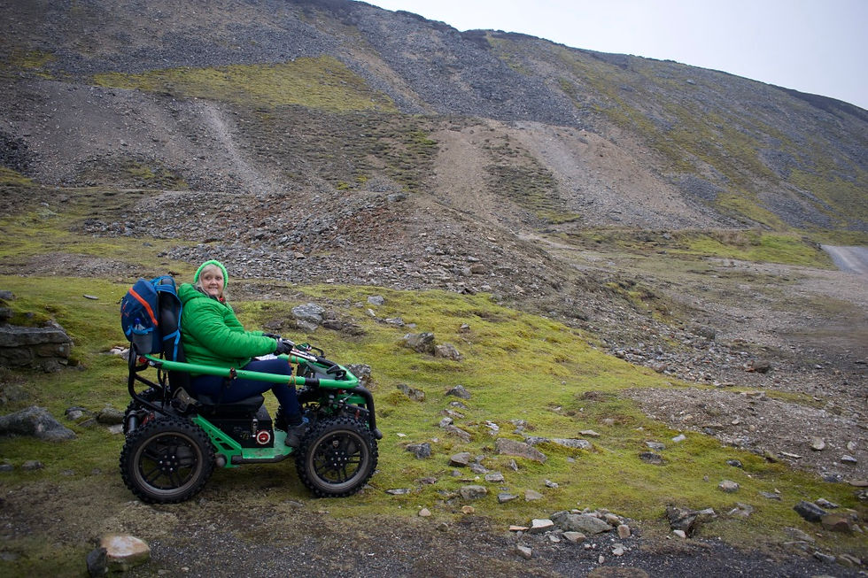 Person in a green jacket sitting on a rugged wheelchair, smiling on a rocky hill with a grassy foreground. Overcast sky.