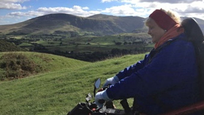 Debbie North in an all-terrain wheelchair on Latrigg, with green hills and the Lake District fells stretching behind her beneath a partly cloudy sky.
