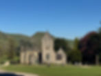 Stone church with a clock on its tower, surrounded by greenery and hills under a clear blue sky. A few people walk nearby. Peaceful mood.