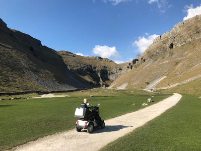 Person on a mobility scooter on a path in a sunny valley. Green grass, rocky hills, and a clear blue sky create a serene atmosphere.