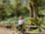 A person in a white shirt sits in a wheelchair by a large tree. A black and white dog walks nearby. Lush greenery surrounds them.