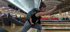 A women in a bowling alley, bowling with her baby sleeping in a structured baby carrier on her back.