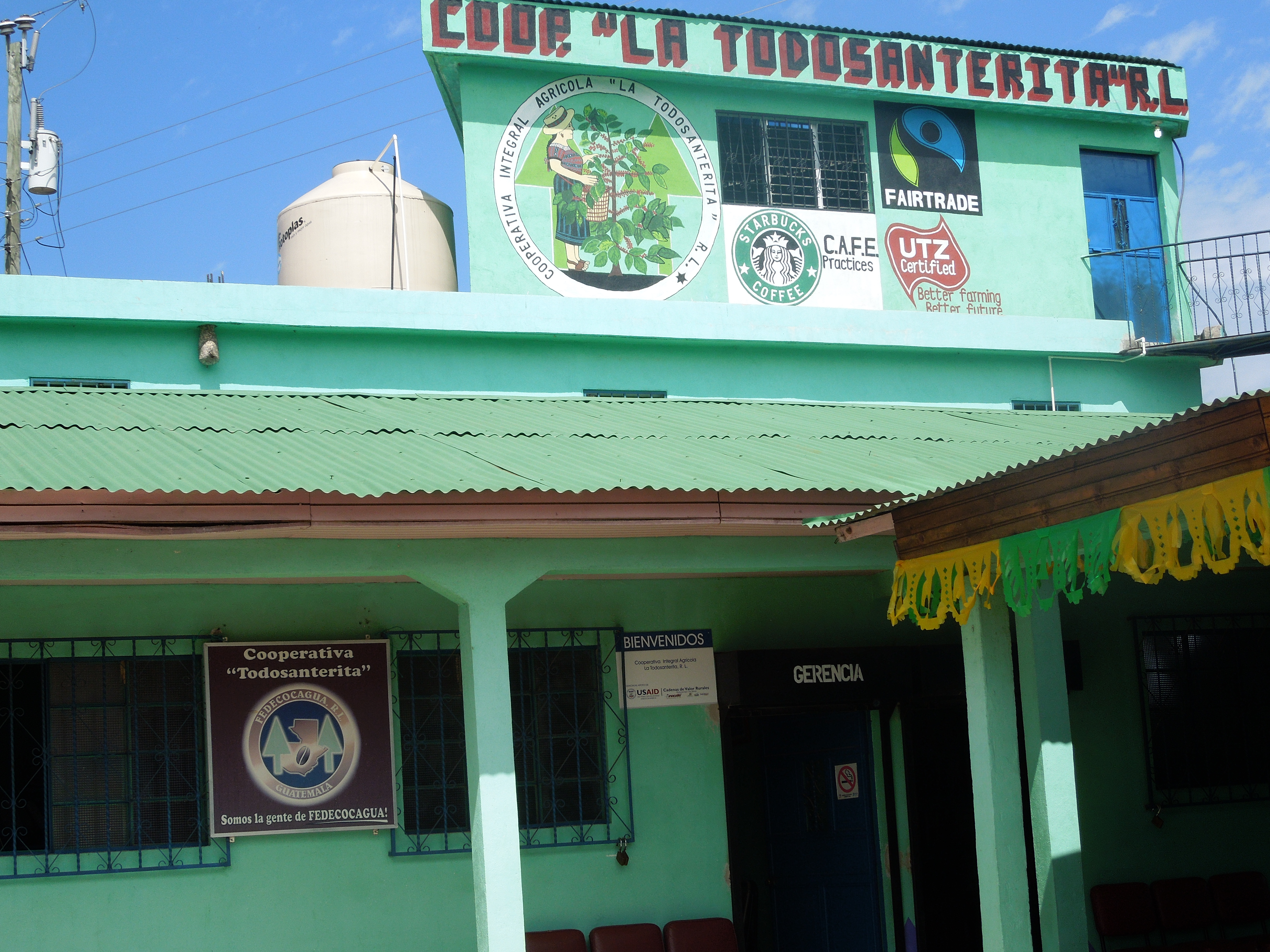 Green building labeled "Cooperativa Todosanterita" with logos for Fair Trade, Starbucks, and UTZ. Festive banners hang above. Sunny day.