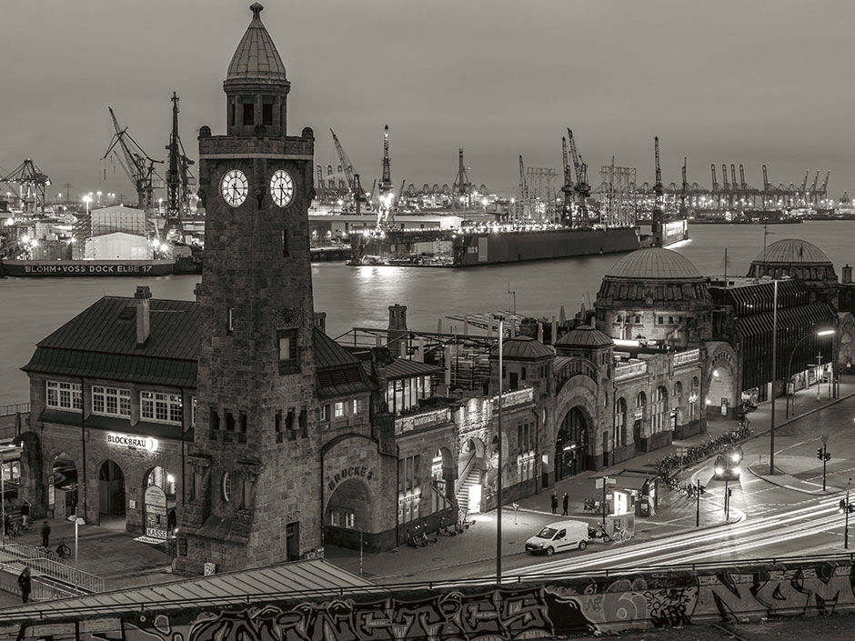 The Landungsbrücken clock tower and harbour terminal in Hamburg, photographed at night in black and white, with the port's industrial cranes visible across the water.