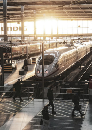 Blick in die Gleishalle des Münchner Bahnhofs mit einem ICE vor dem Sonnenuntergang in der Rückwand aus Glas.