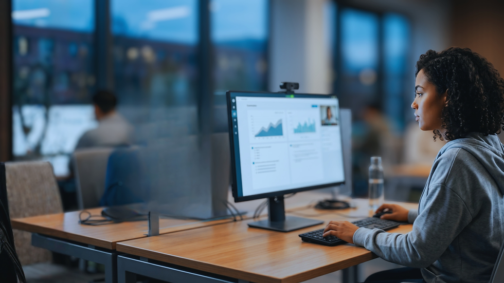 Woman in a grey hoodie works on a computer displaying graphs in a modern office with large windows. Focused atmosphere, blurred background.