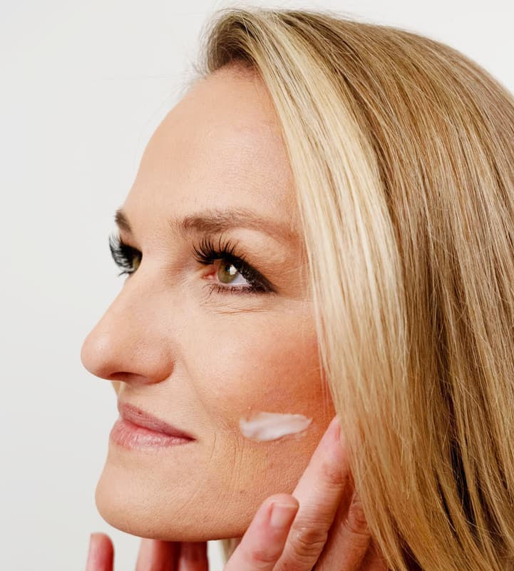 A woman with long blonde hair applying a cream to her cheek, looking upwards.