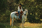 A woman confidently riding a brown horse through a grassy field under a clear blue sky.