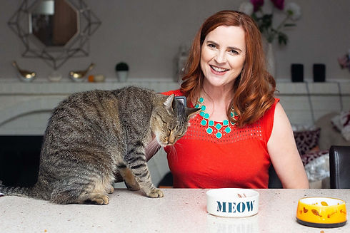 Woman with red hair smiles next to a cat at a table with a bowl labeled "MEOW.