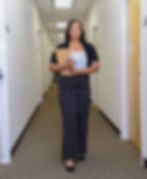 A woman in a light top, black cardigan, and striped pants walks down a hallway holding a clipboard and pen.