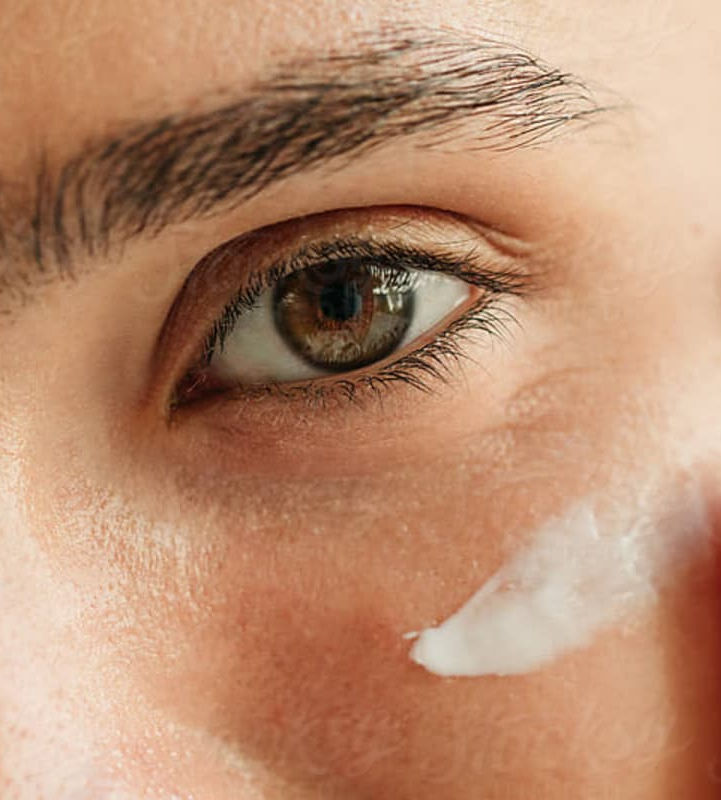 A close-up of an eye area, with a finger applying a small amount of white cream to the skin.