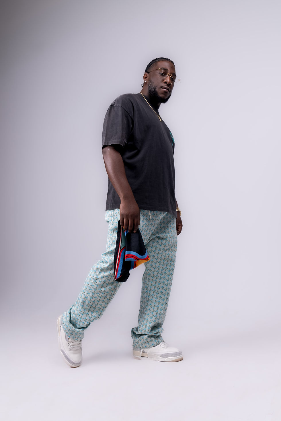 Kincy with dreadlocks, wearing a dark t-shirt, light blue patterned pants, and white sneakers, holding a colorful folded garment.