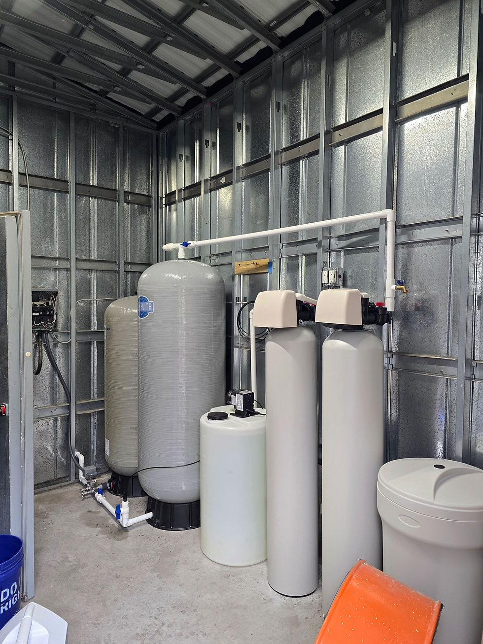 An industrial commercial water treatment system featuring large grey mineral tanks, white softener cylinders, a brine tank, and interconnected white PVC plumbing installed in a room with metal wall framing.