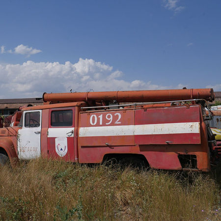 Abandoned ZIL-130 fire trucks in a field, Rusting Soviet fire truck with Cyrillic markings, Soviet ZIL-130AC with ladders and hoses, Graveyard of fire trucks near factory ruins, Soviet fire engines lined up in overgrown grass