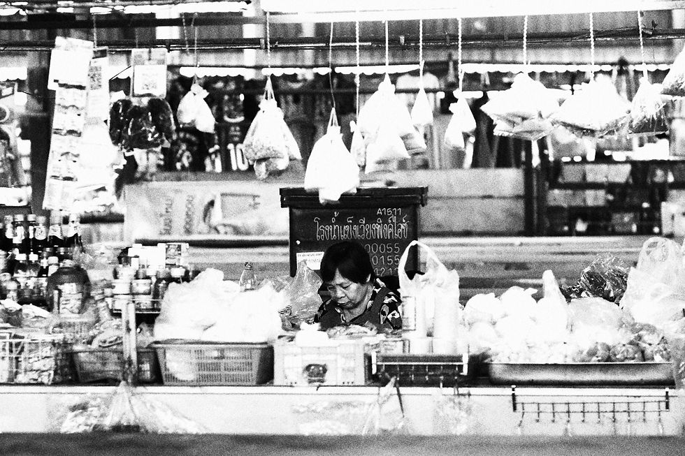 Black and white photo of a market vendor working behind a counter, with rows of bagged goods hanging above her.