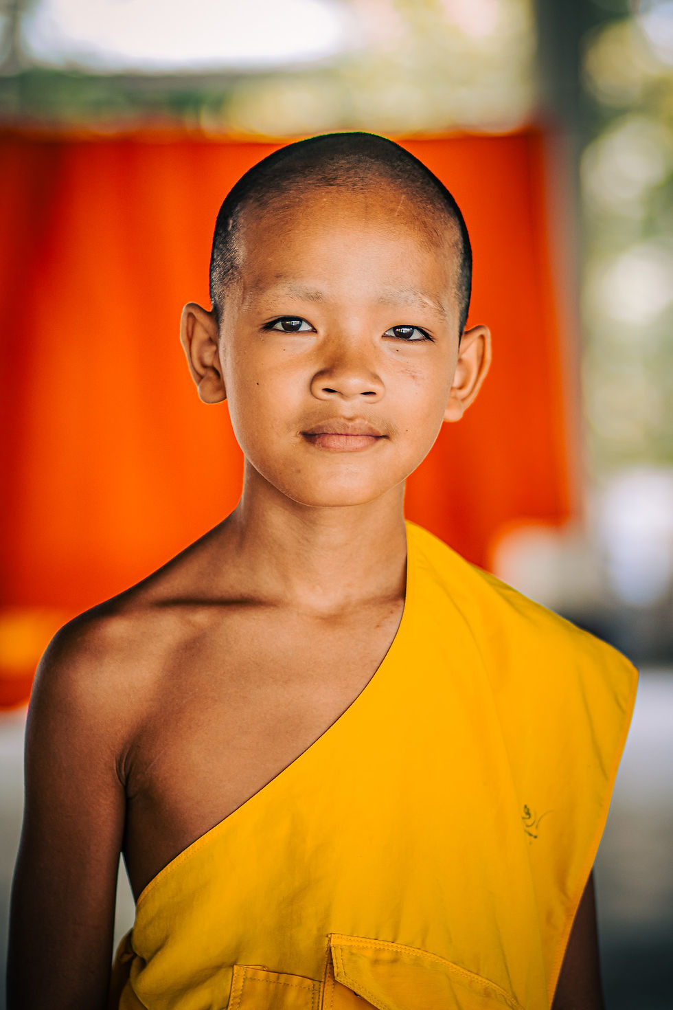 Portrait of a young Thai novice monk in bright yellow robes standing in front of a vivid orange curtain, with soft light illuminating his calm expression.