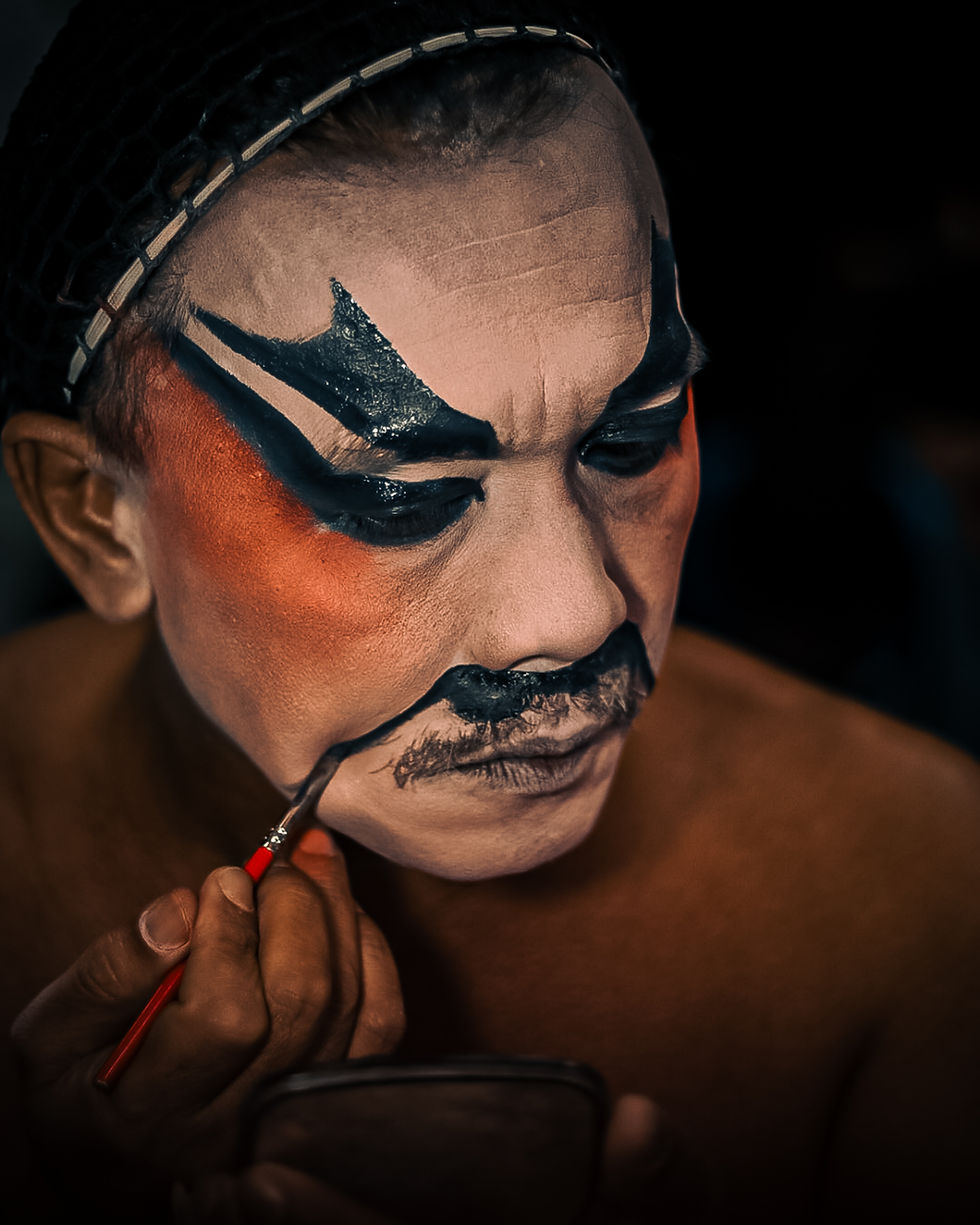 Close-up of a Chinese opera performer painting a black mustache on their heavily stylized face makeup, showing intense concentration and cultural symbolism.