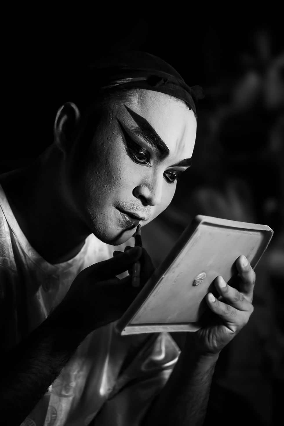 Black and white portrait of a male Chinese opera actor applying lip makeup while looking into a handheld mirror, partially dressed in costume.