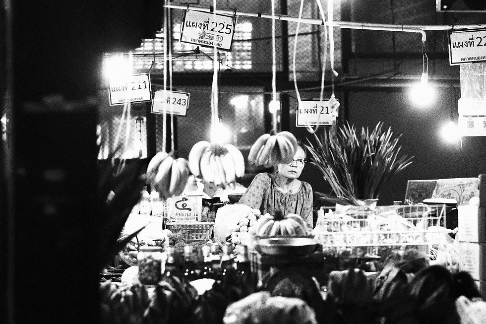 Market vendor framed behind hanging banana bunches and bare lightbulbs in a grainy black and white scene.