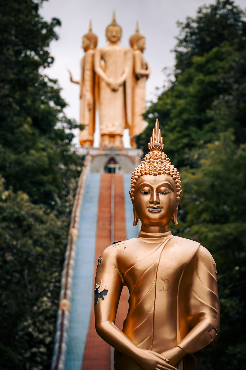 A golden Buddha statue with weathered patches stands in front of a staircase, leading to a monumental four-faced Buddha structure at the top of a forested hill at Wat Doi Sapphanyu in Thailand