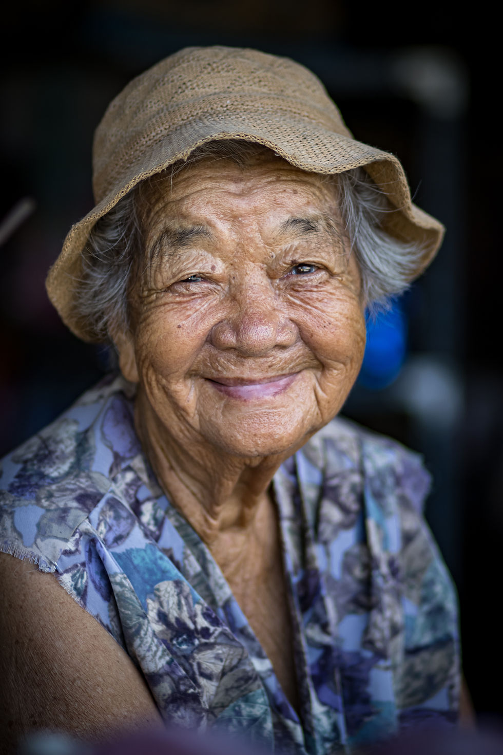 Portrait of an elderly Thai woman in a floral blouse and straw hat, smiling warmly at the camera — taken with permission during a street photography session.