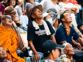 Crowd of Thai people, including a monk and elderly villagers, looking up in anticipation during the Bamboo Rocket Festival in Chiang Mai.