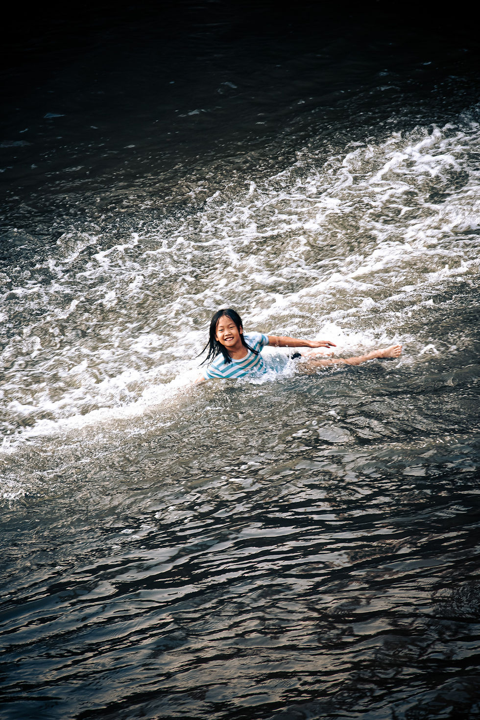Smiling Thai girl lying in the flowing water current at Saraphi bridge Chiang Mai, candid joyful child photography Thailand