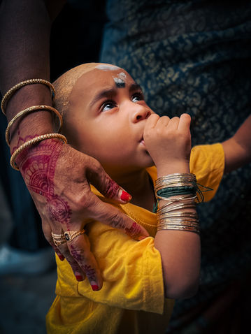 Infant sucking their thumb during a traditional blessing, with henna-stained hands of an elder nearby, at Batu Caves in Malaysia.