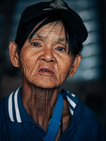 Older Thai woman in a cap, showing strength and resilience in her quiet expression.