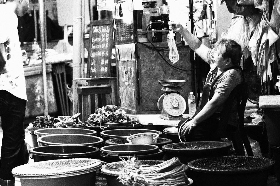 Grainy black and white photo of a fish vendor seated among tubs, holding up a tied plastic bag of fish toward a customer; vintage weighing scale and price board in the background.