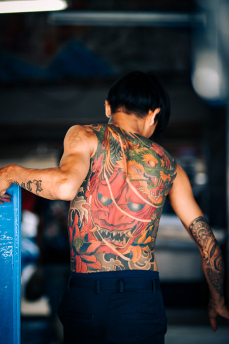 Shirtless Thai man with full back tattoos walking through an urban street scene.