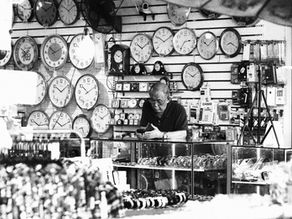Grainy black and white photo of a Chiang Mai clock shop owner at his counter, surrounded by wall clocks, wristwatches, and shop displays.
