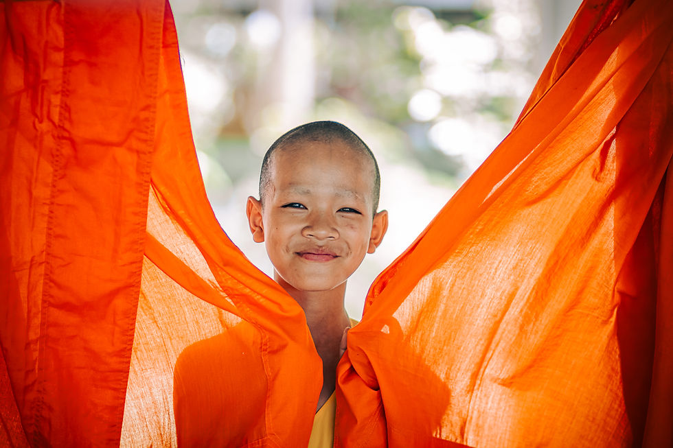 Smiling novice monk playfully stands between two fluttering orange monk robes, capturing a candid moment of joy inside a Thai temple.