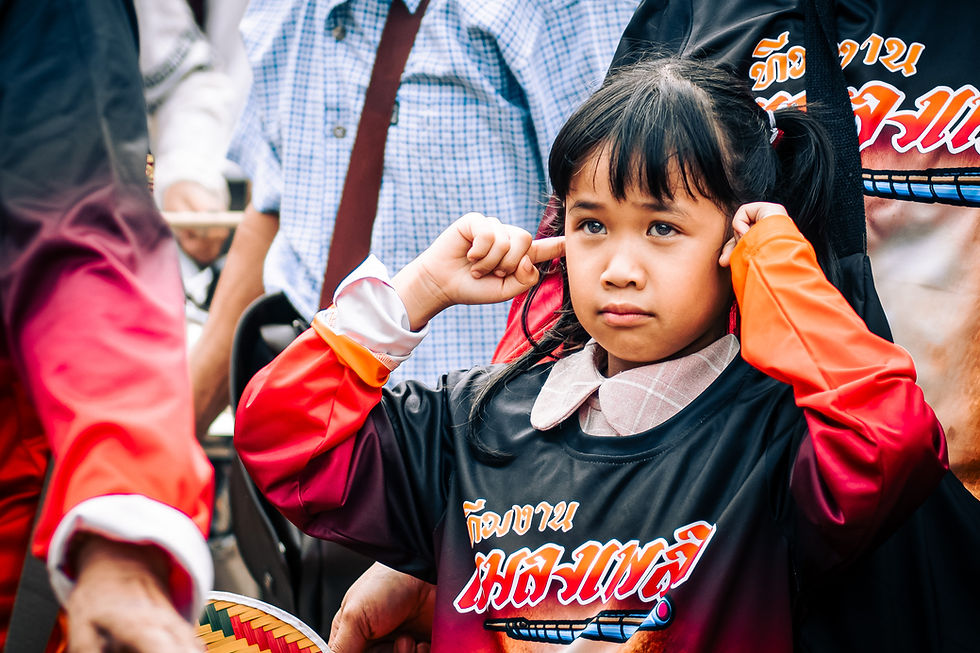 Young Thai girl covering her ears during the Bamboo Rocket Festival in Chiang Mai, dressed in a team shirt and surrounded by adults.