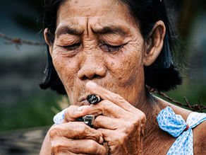 Portrait of an older Thai woman with short black hair, holding a lit cheroot in her mouth, dressed in a blue polka-dot dress, standing near a wire fence
