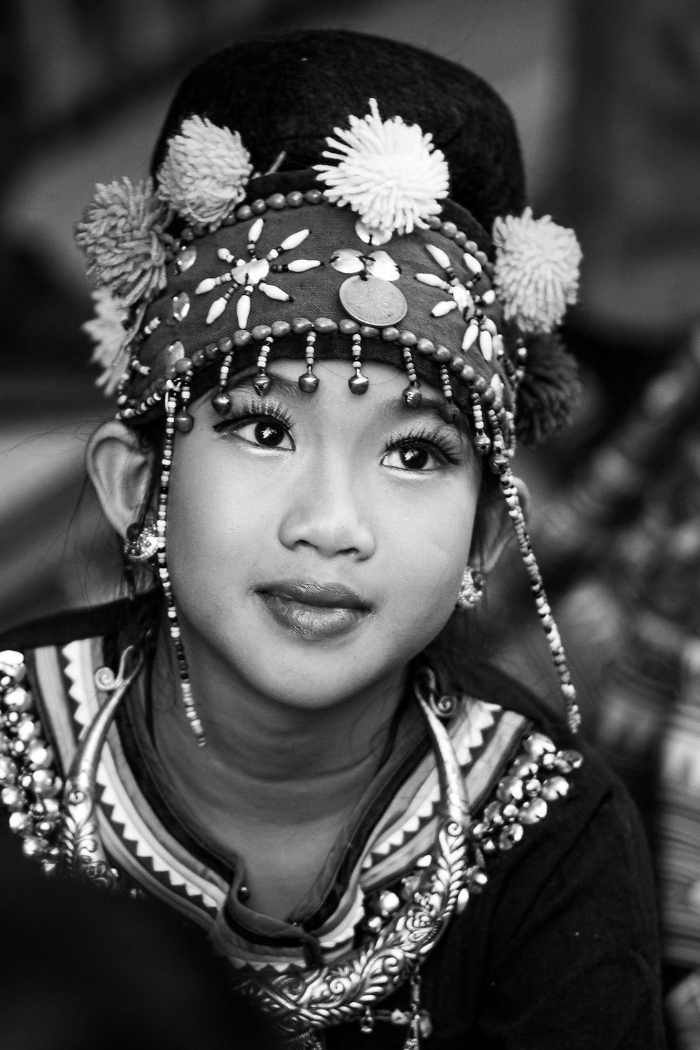 Close-up black and white portrait of a young Thai girl in ceremonial hill tribe attire, looking upward with thoughtful eyes.