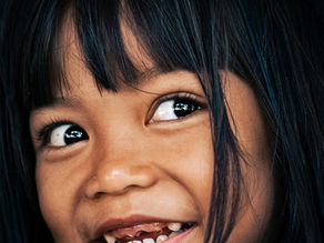 Close-up portrait of a young girl with long black hair and a playful smile, missing front teeth, taken in natural light in Central Java, Indonesia.