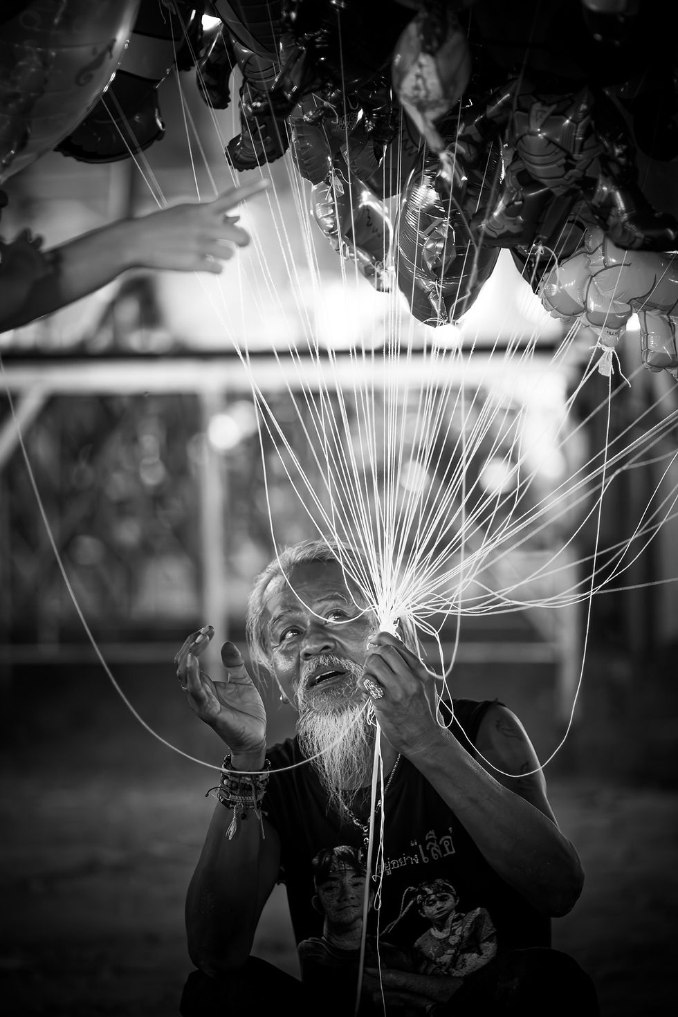 Black and white portrait of an elderly balloon vendor looking up while holding tangled strings, with a hand reaching from above to select a balloon.
