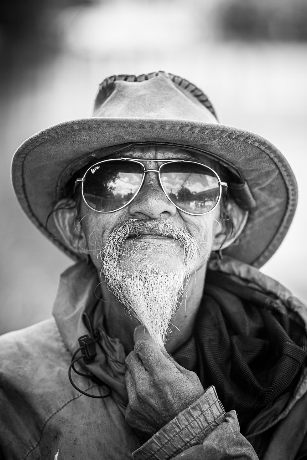 Black and white close-up portrait of an elderly man wearing sunglasses and a sunhat, gently holding his beard with a contemplative expression.
