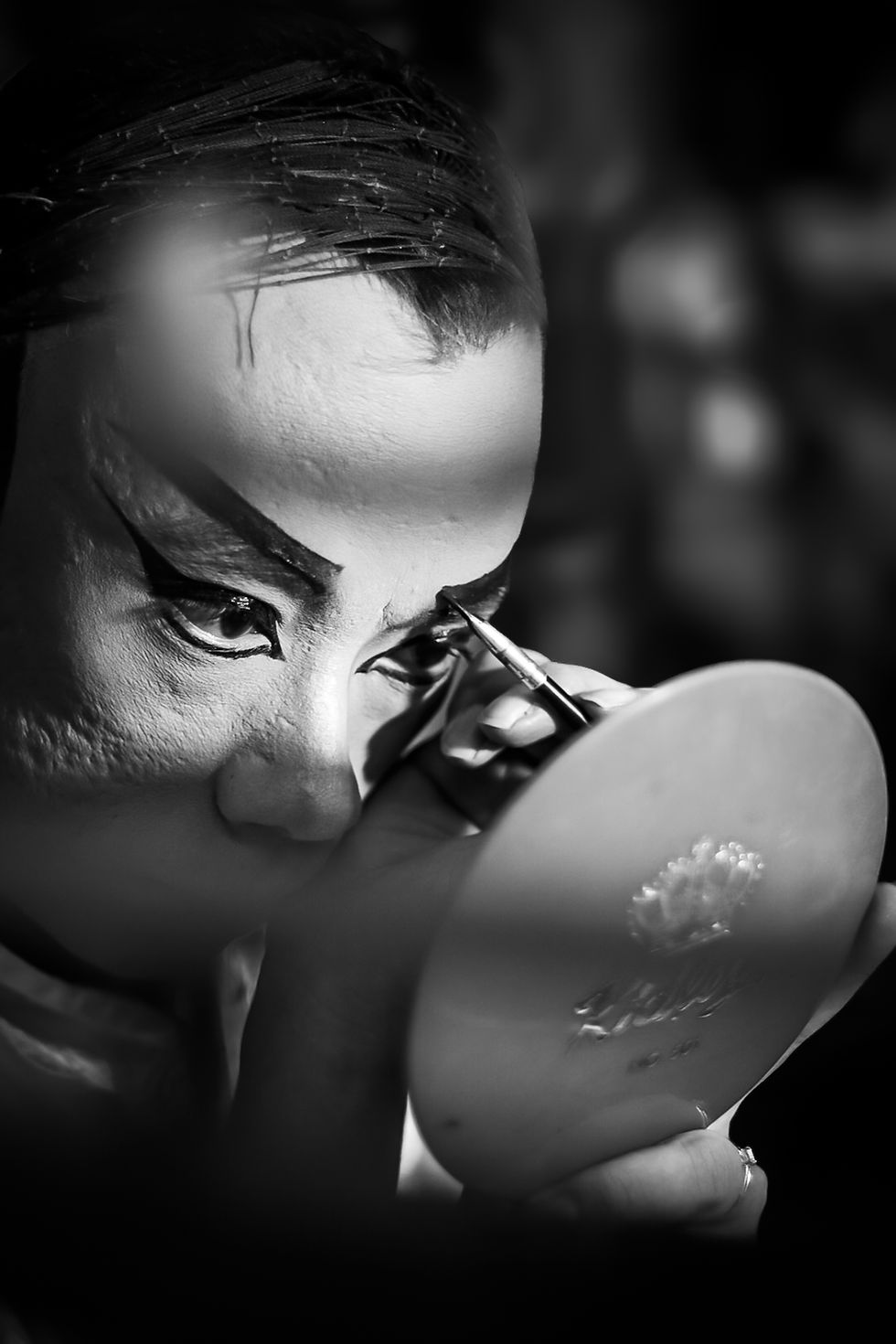 Close-up black and white photograph of a Chinese opera performer applying dramatic eye makeup, with intense focus reflected in their gaze.