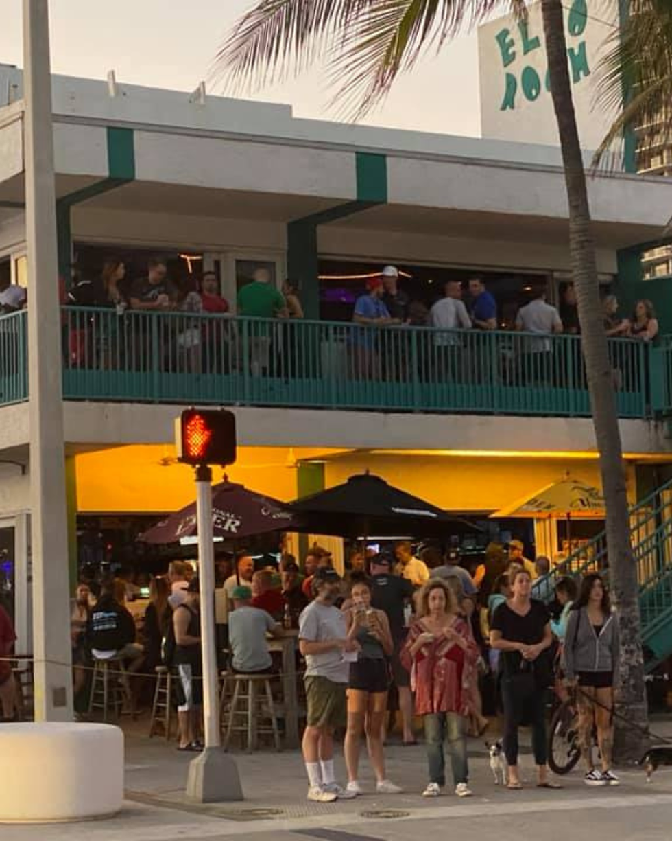 People gather outside a lively bar with outdoor seating, umbrellas, and palm trees. A red light signals pedestrians at sunset. Building text reads "ELBO ROOM."