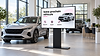 Illustration de mise en situation générée par IA : showroom automobile lumineux avec deux véhicules sans marque et un écran professionnel présentant les rubriques Découvrir, Essayer et Entretenir.