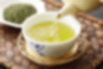 Hot honey green tea pouring from a pot into a white tea cup with indigo accents. Sitting on a woven wood tray with a bowl of loose green tea leaves
