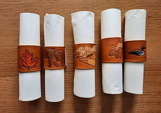 5 leather napkin rings with napkins. Brown napkin rings feature a different design. From left to right designs are: A red Maple Leaf, a dark brown bear, a Cayak on a lake in the moutains, a moose head, and a loon